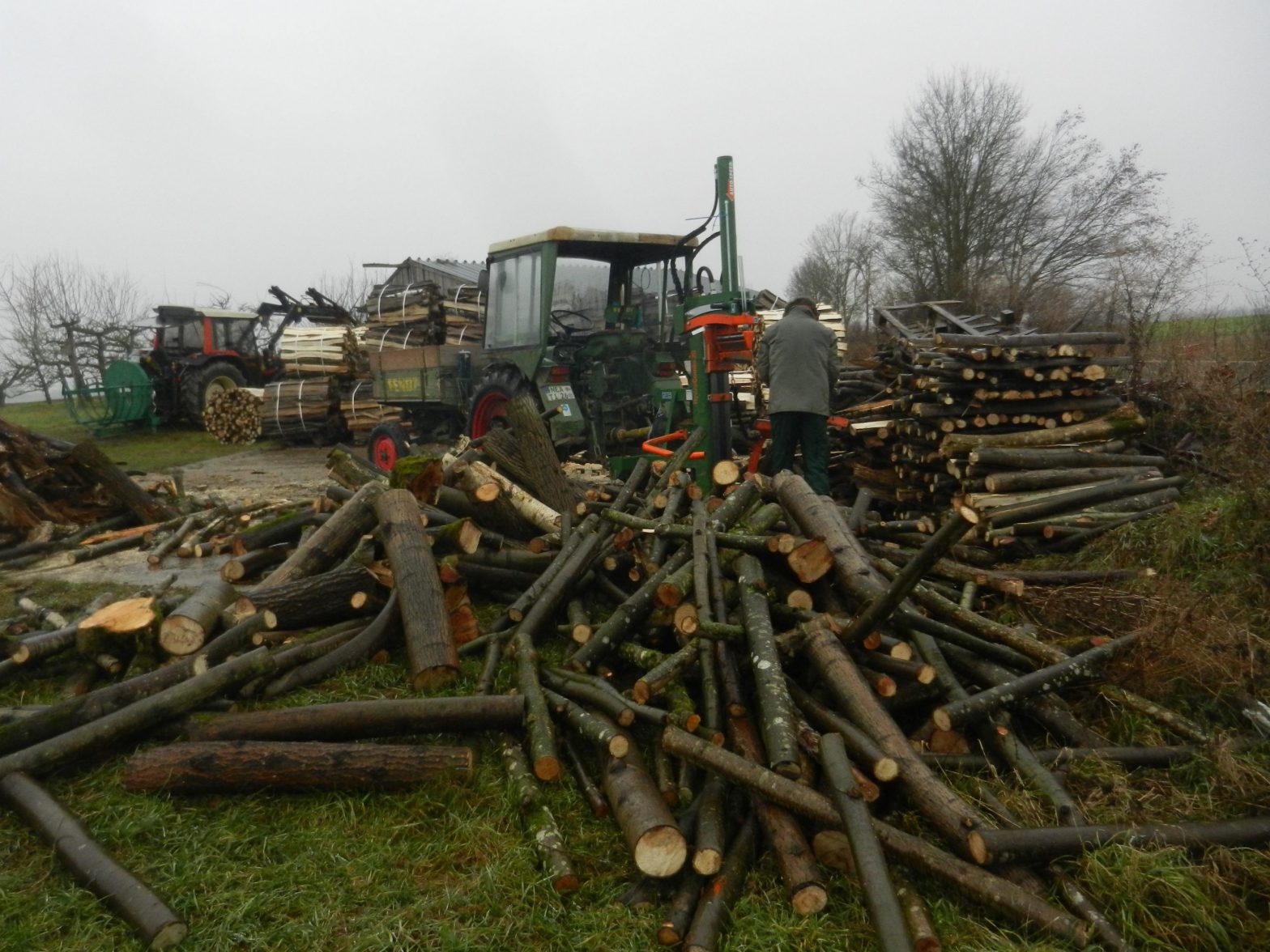 Traktor mit Holzspalter mit arbeitender Person. Haufen mit losem Holz im Vordergrund und Stapel mit fertig gespaltenen Scheiten im Hintergrund