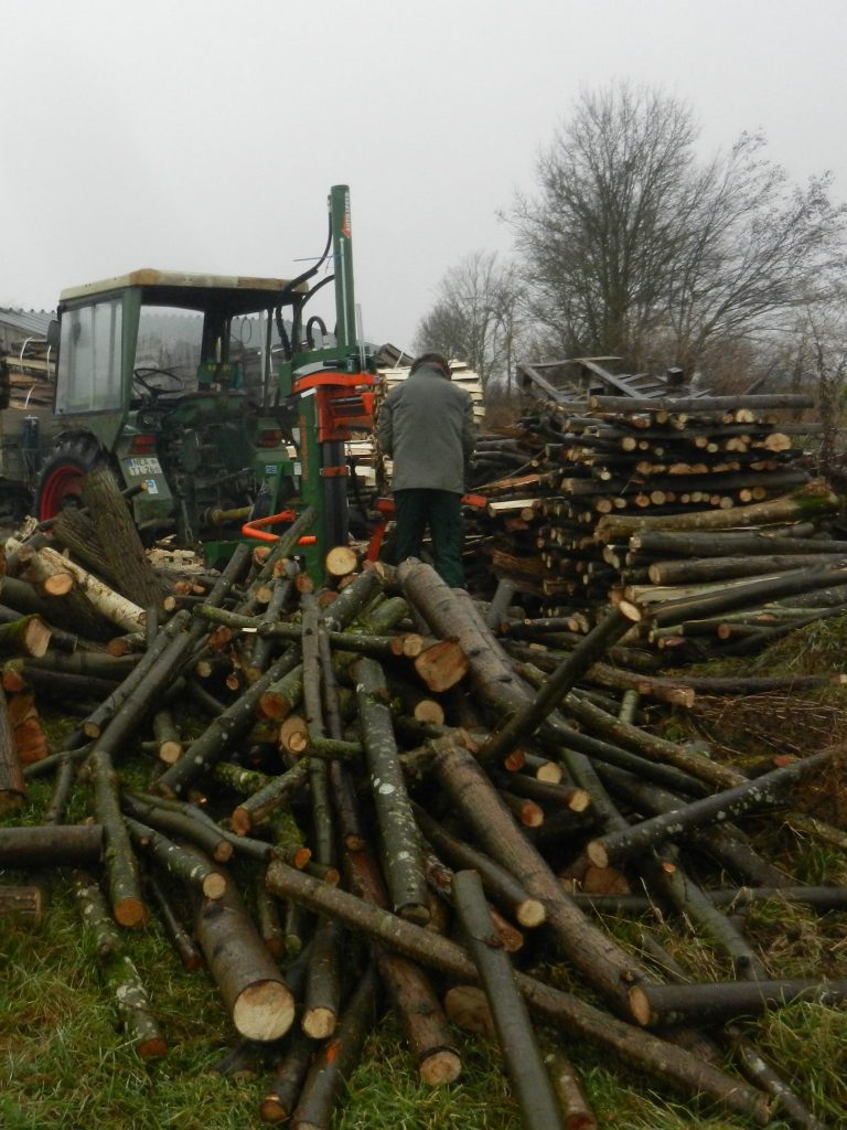 Traktor mit Holzspalter mit arbeitender Person. Haufen mit losem Holz im Vordergrund und Stapel mit fertig gespaltenen Scheiten im Hintergrund